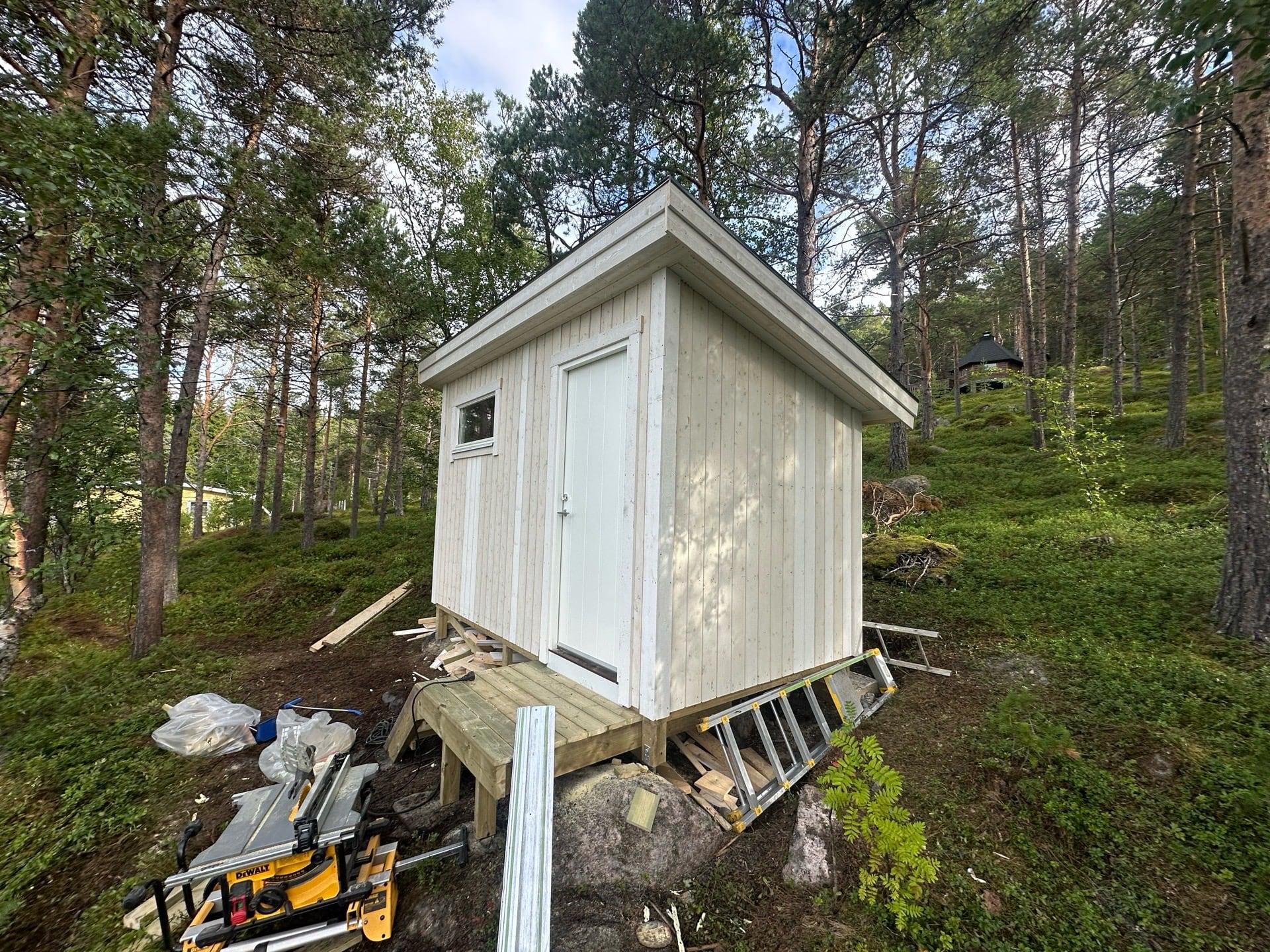Hvit bod på stolper i skogen, verktøy på bakken foran